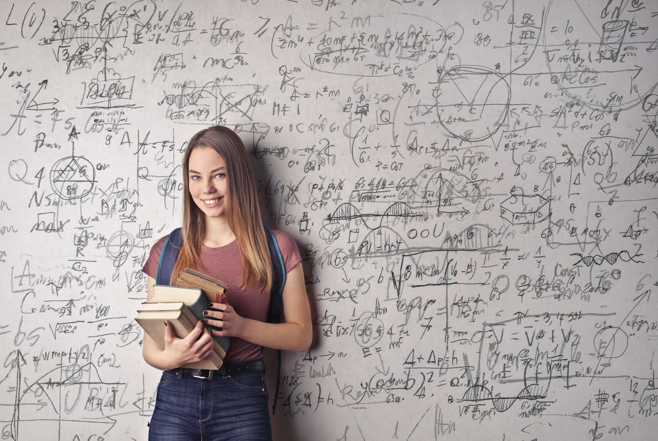 hero-gallery-03 Young woman holding books standing in front of a chalkboard filled with mathematical formulas.