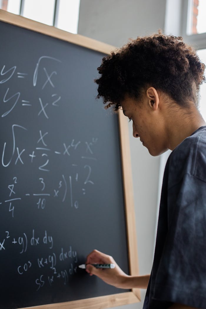 about-img Side view of African American learner making math calculations on blackboard during lesson in classroom